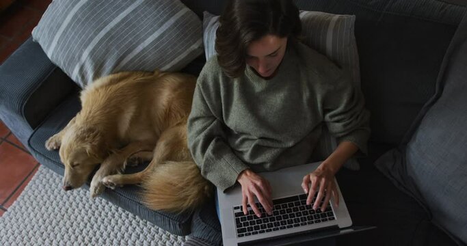 Smiling caucasian woman using laptop working from home with her pet dog on sofa next to her