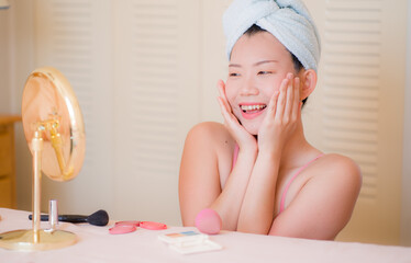 young beautiful and happy Asian Korean woman with towel head wrap applying makeup looking to mirror smiling cheerful preparing for dating in beauty fashion concept