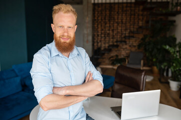 Successful redhead man wearing smart casual shirt stands in confident pose with arms crossed, confident red haired male entrepreneur in home office looks at the camera