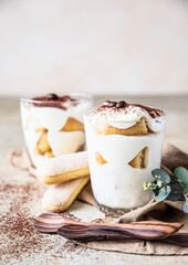 Traditional Italian dessert tiramisu in glass, light stone background. Coffee flavored portion dessert made of ladyfingers and mascarpone decorated with cocoa powder.
