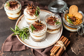 Layered dessert in glass jar with cookie crumble and whipped cream decorated with rosemary and anise, dark brown background. No bake cheesecake, trifle or pudding.