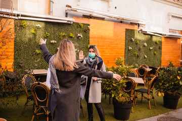 A beautiful young blonde woman with a medical face mask greets her friend in the center as she motions for her to stop. Shopping during the COVID-19 coronavirus epidemic. Social distance