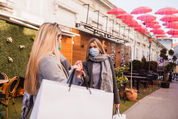 Two beautiful young Caucasian women with a face mask greet each other on the street. Shopping during the COVID-19 coronavirus pandemic