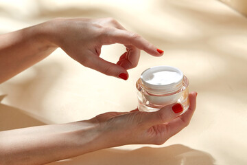 beauty product, cosmetics and people concept - female hand holding jar of moisturizer on beige...