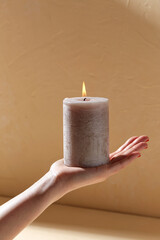 aromatherapy, coziness and people concept - female hand holding burning aroma candle on palm over beige background with shadows