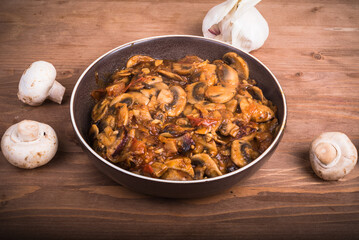 Portion of sautéed champignons with tomatoes in a plate on the table with raw mushrooms