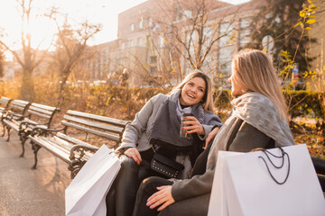 Two influencer girls with the phone sitting on the bench after shopping - Happy friends talking about  sales - Concepts about lifestyle, shopping, technology and friendship