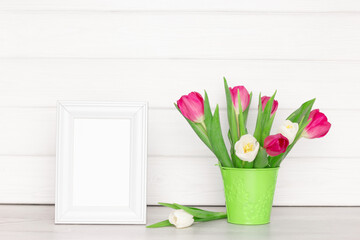Empty white photo frame with tulip flower bouquet on table near wooden board wall. Light rustic interior. Bright spring mock-up background