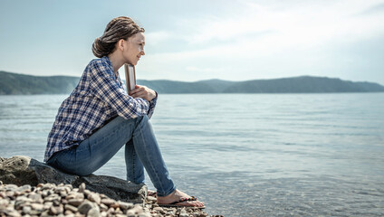 Woman is sitting on the stony coast of a large pond with a book in her hands and enjoying solitude and nature. Concept of a dreamy mood, lifestyle and relaxation from the hustle and bustle of the city