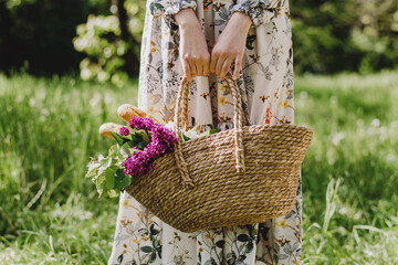 Female holding picnic basket with food and bouquet of lilacs outdoors.