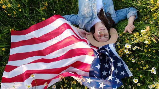 July fourth concept. Top view portrait of cheerful young woman lying on green grass with usa United States flag under sunny day. Smiling free proud independent patriotic happy girl feeling freedom