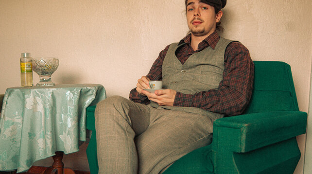 man in vintage suit is sitting on sofa and holding coffee cup. an old house. old colors. vintage background