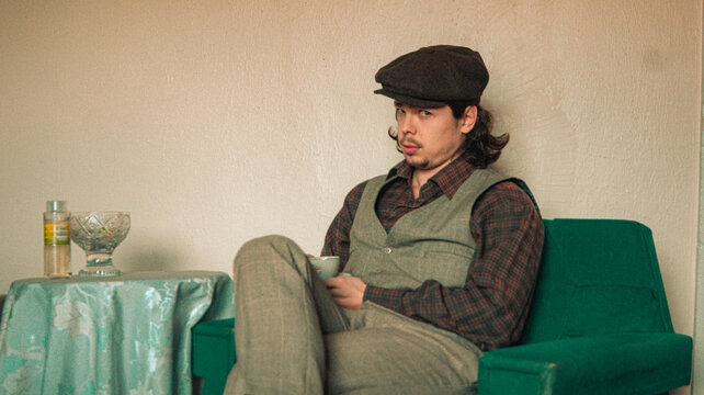 man in vintage suit is sitting on sofa and holding coffee cup. an old house. old colors. vintage background