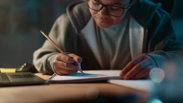 Young Teenage Multiethnic Black Girl Writing Down Homework in a Notebook with a Pencil, Using Laptop Computer in a Dark Cozy Room at Home. She's Browsing Educational School Research Online.