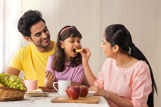 A HAPPY MOTHER FEEDING HER DAUGHTER WITH HUSBAND SITTING NEAR	