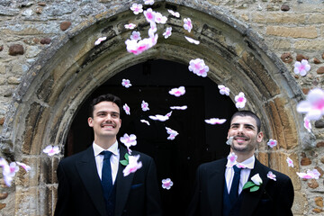Smiling gay grooms leave church after getting married with confetti falling around them.
