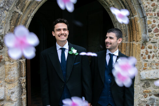 Smiling gay grooms leave church after getting married with confetti falling around them.