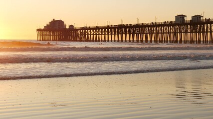 Wooden pier on piles, silhouette at sunset, California USA, Oceanside. Waterfront resort, pacific ocean tide, tropical beach. Summertime coastline vacations atmosphere. Sunny sea waves at sundown.