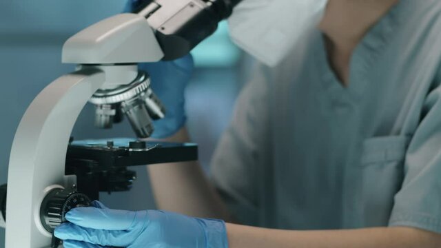 Tilt up shot of female scientist in protective gloves, mask, glasses and hat looking through microscope while doing research in lab