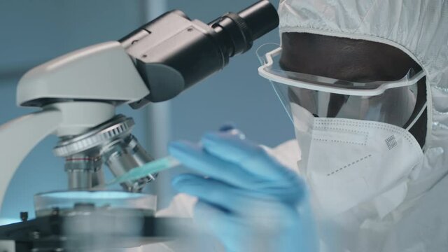 Tilt down shot of Afro-American male lab worker in protective suit, gloves, mask and glasses pouring blue liquid from pipette under microscope while conducting research during coronavirus pandemic