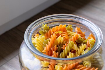 A portrait of a glass pot, jar or bowl full of spirelli in three different colors, orange, yellow and green. The uncooked tricolore italian pasta variant is swirl shaped.
