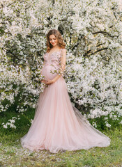 Obraz na płótnie Canvas Young beautiful happy joyful pregnant woman in light pink long evening dress posing. background of blooming trees. Garden green leaves white flowers. Girl model. Elegance pregnancy spring fresh nature