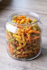 A portrait of a glass pot, jar or bowl full of spirelli in three different colors. The uncooked tricolore italian pasta variant is yellow, orange and green and is swirl shaped.