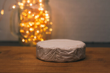 A whole head of brie or camembert cheese with white mold on a wooden cutting board. Christmas lights in a glass jar. Preparation of a cheese plate. Snack for the holiday or party - obrazy, fototapety, plakaty