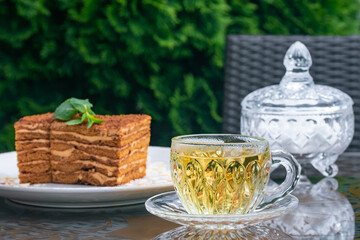 Crystal cup with mint tea, sugar bowl and medovik cake on table in summer cafe. Tea drinking concept, British culture. Still life. Lifestyle. Traditional Russian cuisine, dessert.