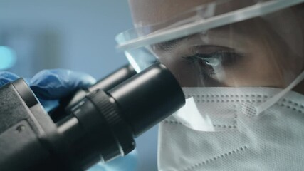Close up shot of female scientist in protective uniform, mask and glasses looking through microscope while doing lab research during covid- outbreak - Powered by Adobe