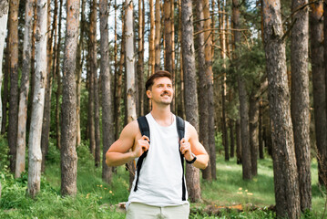 Man hiking outdoors. Handsome smiling young man with backpack standing in pine forest and looking to the side. Happy traveler having a rest in nature, sunny summer day. Hike, travel, trekking concept