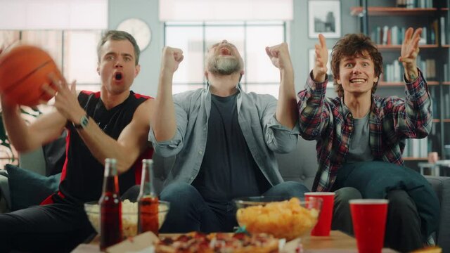 At Home Three Joyful Basketball Fans Sitting on a Couch Watch Game on TV, Celebrate Victory when Sports Team Wins Championship. Group of Friends Cheer When Favourites Play. Wide Portrait