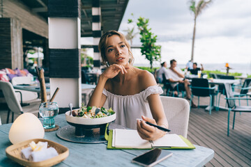 Young female sitting at table in cafe and thinking in summer day