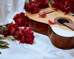 An acoustic wooden guitar lies on a white sheet surrounded by red peonies.