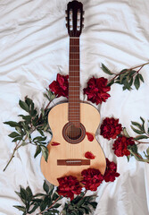 An acoustic wooden guitar lies on a white sheet surrounded by red peonies.