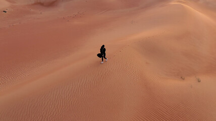 Fototapeta na wymiar AERIAL. Camera following woman in traditional Emirati dress walking in a desert in strog wind and sunset.
