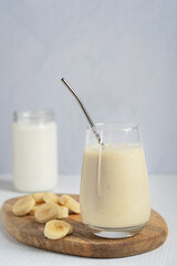 Popular lassi drink or milkshake made of ripe sweet bananas blended with milk served in drinking glass with zero waste metal straw on cutting board on white wooden table for breakfast. Vertical image
