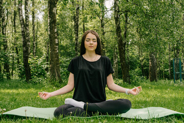 a girl in a black T-shirt and leggings sits in a lotus position on a sports mat in greenery