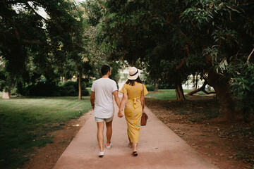 A photo from behind of a brunette girl in a yellow dress and her boyfriend who are walking on a...