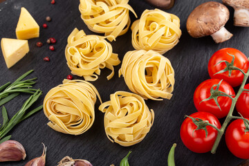 Raw pasta tagliatelle with fresh tomatoes, peppers, mushrooms,asparagus beans, spices and cheese on black table.