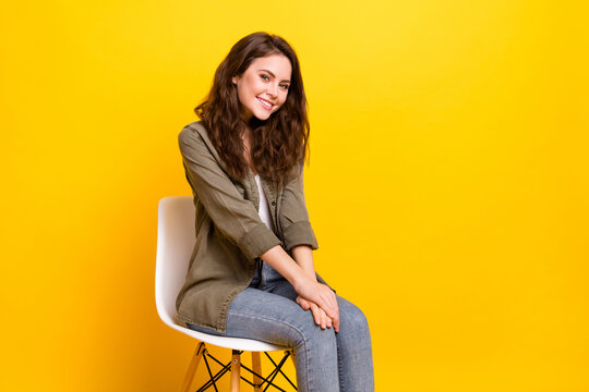 Portrait of pretty shy cheerful girl sitting on chair expecting meeting isolated over vibrant yellow color background