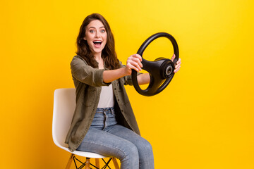 Portrait of attractive cheerful girl sitting on chair holding steering wheel riding having fun isolated over bright yellow color background
