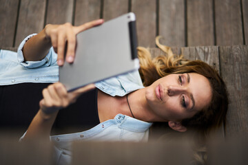 Millennial hipster girl lying outdoors using modern touch pad for online networking in social media, attractive female blogger checking settings of digital tablet while browsing text publication