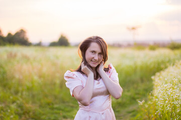 Beautiful portret of happy woman with dark hair standing in the wildflowers field in the long pink dress and sensually touching her face on a warm summer evening. Joyfull summer walking. 