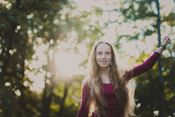 A portrait of a beautiful smiling woman with long blond hair in a purple blouse standing in the park, raising her left hand up. Close up photo of a girl with back sunlight on the nature