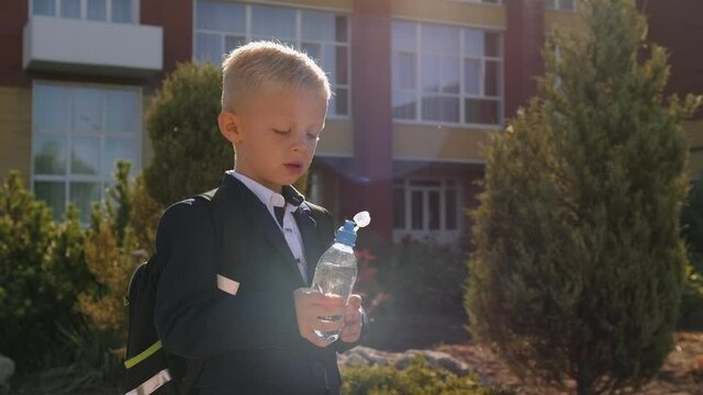 A small schoolboy with a backpack drinks water from a bottle in the schoolyard.