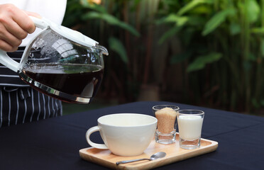 The waiter's hand holds a coffee pot about to pour the coffee onto the cup on the table covered with black cloth. Restaurant atmosphere in the garden with blurred green leaves background.