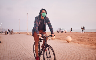 Hombre Joven latino maneja bicicleta durante un bello atardecer.. Joven feliz  con mascarilla en la playa