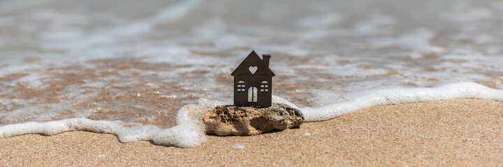 Miniature house on natural sea stone stands on sandy beach, washed by wave. Insurance, rental, sale...