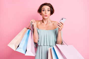 Photo of young girl amazed shocked surprised use cellphone hold bags shopper isolated over pink...